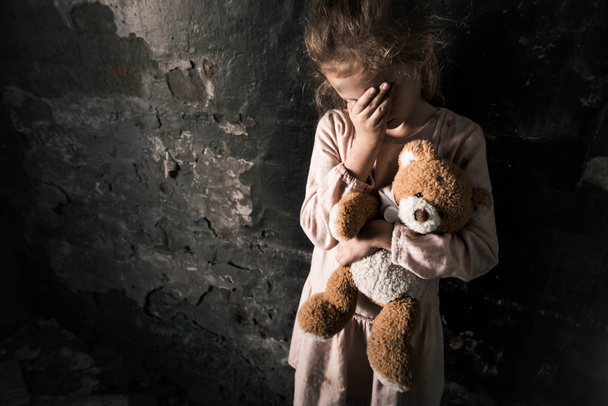 aufgebrachtes Kind berührt Gesicht, während es Teddybär in schmutzigem Raum hält, postapokalyptisches Konzept - Foto, Bild