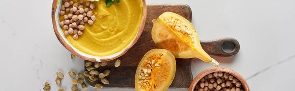 top view of autumnal mashed pumpkin soup in bowl on wooden cutting board near pumpkin, seeds and chickpea on marble surface - Photo, Image