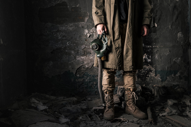 vista recortada del hombre sosteniendo máscara de gas mientras está de pie cerca de la vieja pared en habitación abandonada, concepto post apocalíptico
 - Foto, Imagen