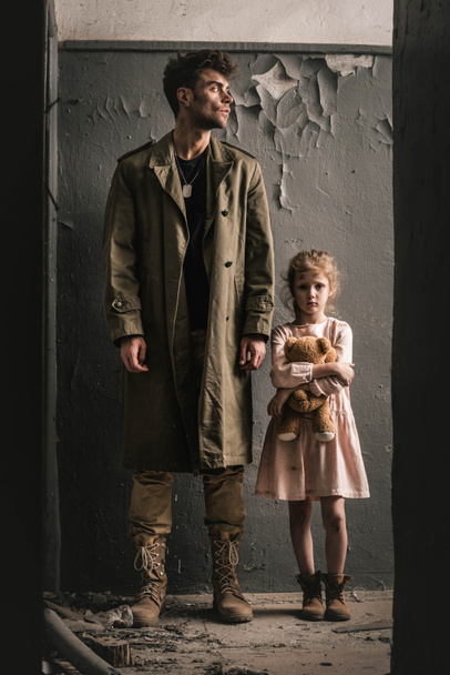 jóképű férfi áll gyerek mellett plüssmackó közel viharvert fal, poszt apokaliptikus koncepció - Fotó, kép