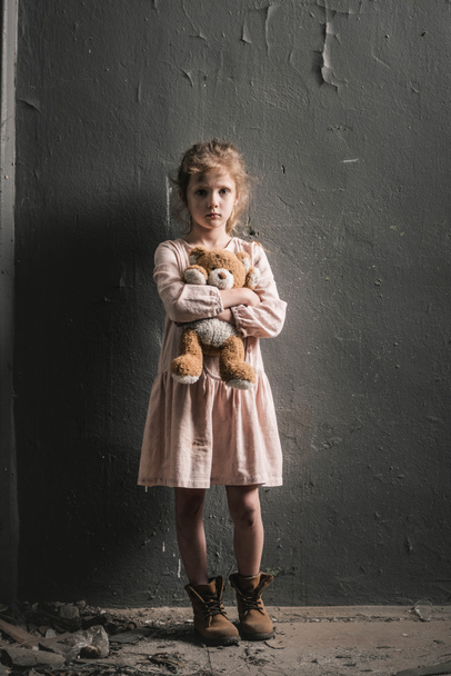 upset kid looking at camera while standing with soft toy near wall, post apocalyptic concept - Photo, Image