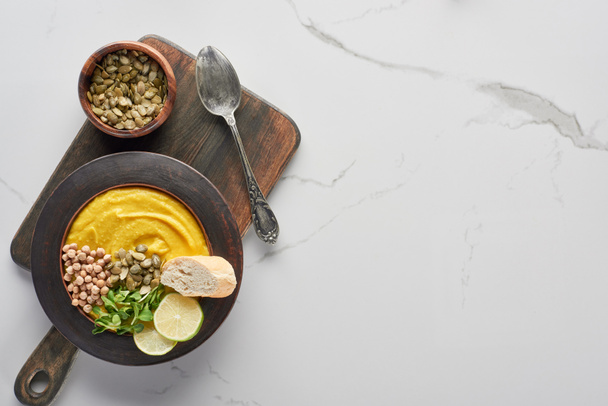 top view of autumnal mashed pumpkin soup in bowl on wooden cutting board with spoon and seeds on marble surface - Zdjęcie, obraz