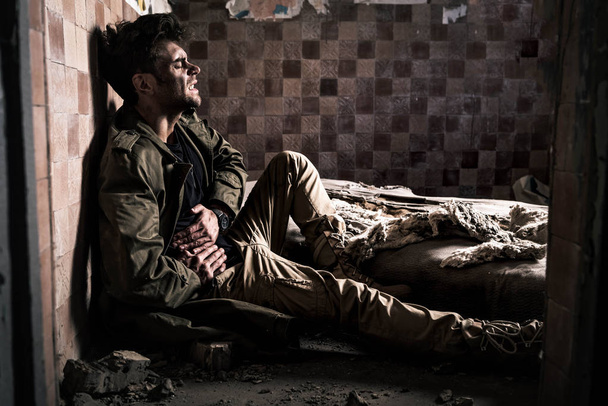 side view of man with dirty face having stomach ache while sitting on floor, post apocalyptic concept - Photo, Image