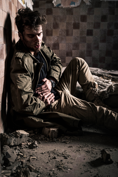 мужчина с грязным лицом, страдающий от боли, сидя на полу, постафокалиптическая концепция
 - Фото, изображение