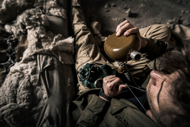 top view of man holding flask while sitting in abandoned building, post apocalyptic concept - Photo, Image
