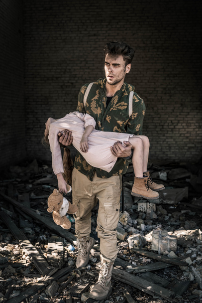 upset man holding in arms kid with teddy bear near abandoned building, post apocalyptic concept - Photo, Image