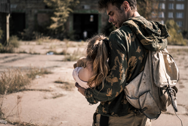 bel homme avec sac à dos tenant dans les bras enfant à l'extérieur, concept post apocalyptique
 - Photo, image