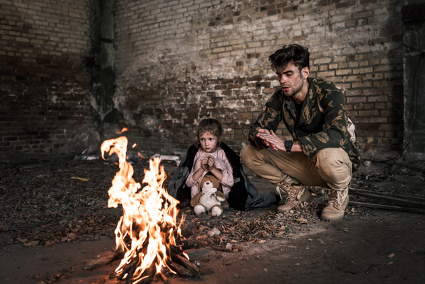 selective focus of man and child sitting near bonfire, post apocalyptic concept - Photo, Image
