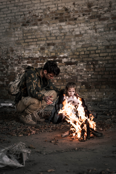 selectieve focus van de man die een jasje draagt op het kind terwijl hij bij het kampvuur zit, post apocalyptisch concept - Foto, afbeelding