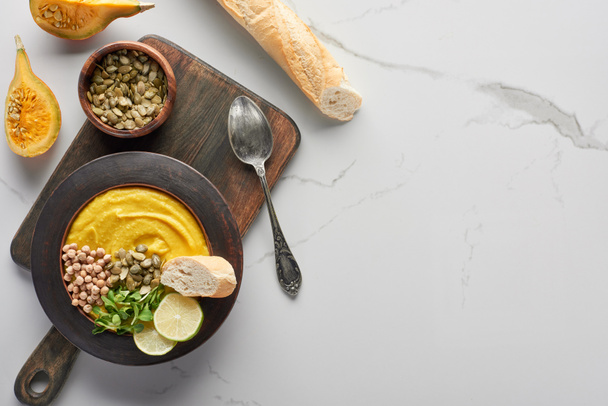 top view of autumnal mashed pumpkin soup on wooden cutting board near silver spoon and baguette on marble surface - Fotografie, Obrázek
