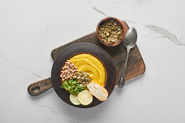 top view of autumnal mashed pumpkin soup on wooden cutting board with seeds and spoon on marble surface - Foto, Imagem