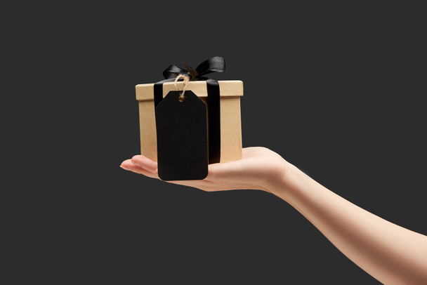 cropped view of woman holding gift box with black blank label in hand isolated on black - Photo, Image