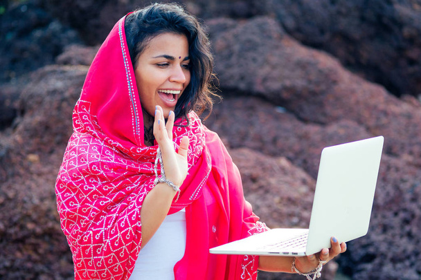 カフェに座っている魅力的なインドのアジアの女性フリーランサー。インドの赤いスタイリッシュなサリーサリーの女性は、ラップトップで作業し、スマートフォンで話して笑顔.リモート仕事の夢の仕事は楽園の海岸で - 写真・画像