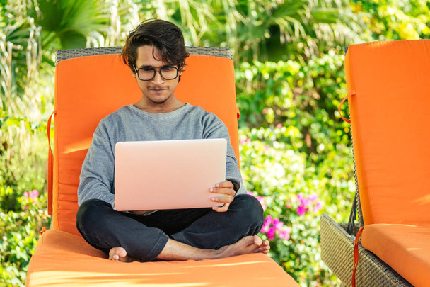 remote work and freelancing.young indian man sitting orange lounger deck chair with laptop on beach.freelancer surfing the internet.freelance dark skinned stylish guy summer vacation tropical paradise - Photo, Image