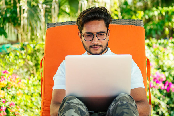 remote work and freelancing.young indian man sitting orange lounger deck chair with laptop on beach.freelancer surfing the internet.freelance dark skinned stylish guy summer vacation tropical paradise - Zdjęcie, obraz
