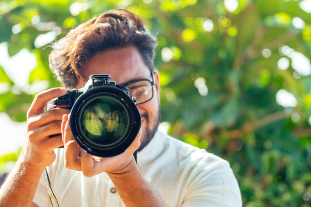 handsome and confident indian man photographer with a large professional camera taking pictures photo shooting on the beach.photo session on summer holiday on the background of green tropical trees - Foto, Imagem