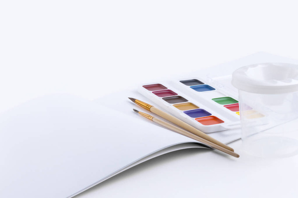 Beautiful composition of bright watercolor paints, two brushes, a container for washing brushes and an open album for drawing with blank pages on a white background - Fotografie, Obrázek