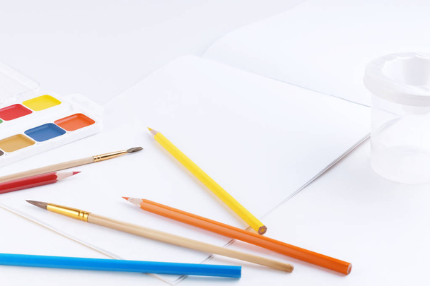 Vista lateral de um álbum de desenho aberto com páginas em branco, tintas aquarelas brilhantes, pincéis de tinta e lápis coloridos em uma mesa branca
 - Foto, Imagem