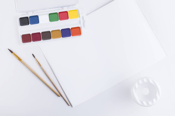 Top view of a beautiful composition of bright multi-colored watercolor paints, brushes for painting, a container for washing brushes and an open album with blank pages for text or a picture - 写真・画像