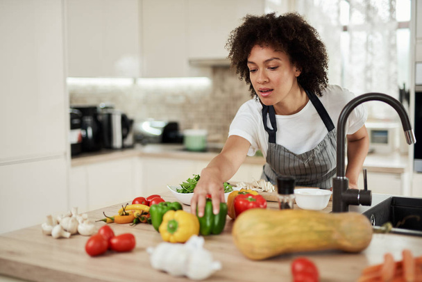 Beautiful mixed race housewife reaching for green pepper while standing in kitchen. Dinner preparation.B. - Foto, imagen