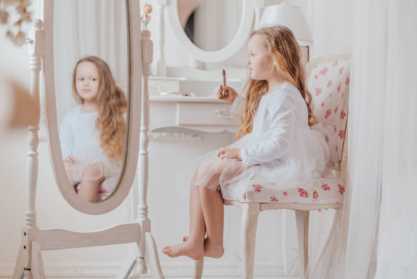 Nahaufnahme Porträt des kleinen Mädchens mit Lippenstift, das allein auf dem Stuhl neben dem großen Spiegel im weißen Raum sitzt - Foto, Bild