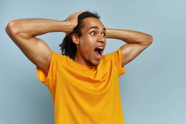 Horizontal portrait of man with dark skin and Afro hairstyle screams in despair, opens mouth widely, being in panic. Frustrated mixed race man poses against blue studio background with copy space. - Foto, Bild