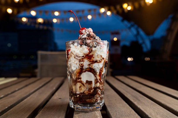 Warstwowy deser lodowy z bitą śmietaną, sosem czekoladowym i lodami waniliowymi na letnim tarasie ze światłami na tle - Zdjęcie, obraz