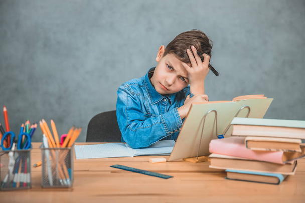 Little tired schoolboy is sitting at the desk, holding hand on the forehead, not knowing what to write, thinking. - Foto, Bild
