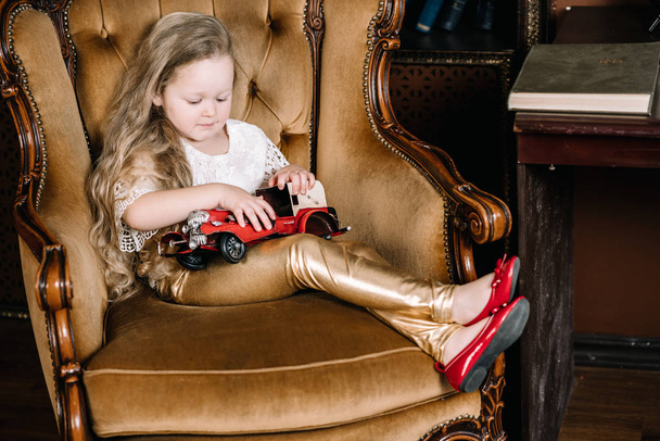 Niña sentada en una silla dorada retro y jugando con un juguete vintage de coche rojo
 - Foto, imagen