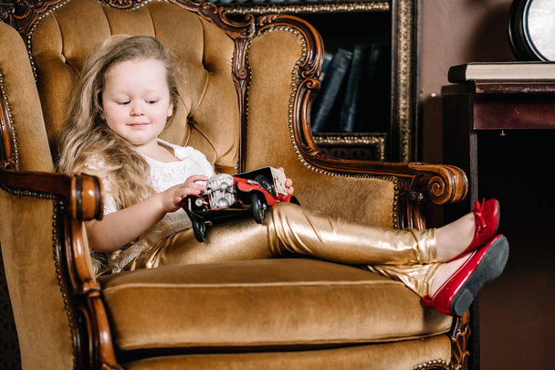 Μικρό μωρό κορίτσι κάθεται σε ρετρό χρυσή καρέκλα και παίζοντας με κόκκινο αυτοκίνητο vintage παιχνίδι - Φωτογραφία, εικόνα