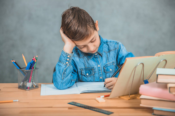 Young concentrated school kid in blue jeans t-shirt writing sitting at table full of books. - Foto, Imagem