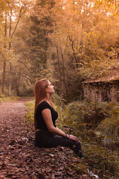 Boční pohled na snící zrzavou ženu sedící v podzimním lese a řece na pozadí při západu slunce. Smutná dívka v depresi, zamyšlená a zamyšlená. Koncept životního stylu. - Fotografie, Obrázek