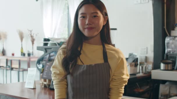 Retrato joven mujer asiática barista sintiéndose feliz sonriendo en la cafetería urbana. dueño de la pequeña empresa coreana chica en delantal relajarse sonrisa dentada mirando a la cámara de pie en el mostrador en la cafetería. - Imágenes, Vídeo