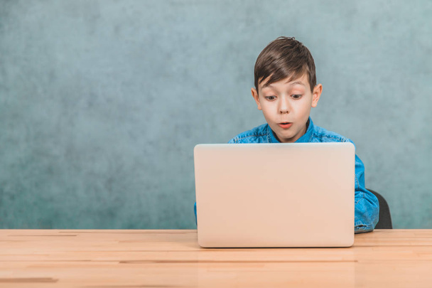 Cute little schoolboy sitting at the desk, surprised by something he has seen in his laptop. - Fotó, kép