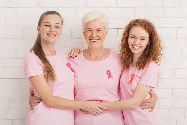 drei Frauen in Brustkrebs-T-Shirts umarmen sich vor weißer Wand - Foto, Bild