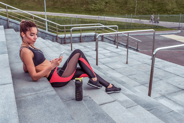 Beautiful girl in sportswear, in summer in city resting after hard training in fresh air, listening to music on headphones, watching video podcast, free space for motivation text. - Фото, изображение