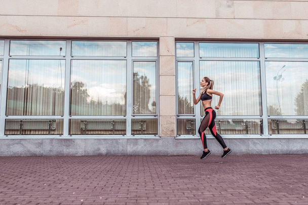 Girl athlete runs background of glass windows, in summer in city on morning run, free space for text motivation fitness workout. Modern lifestyle of youth. Sportswear Leggings. Top. Sneakers. - Фото, зображення
