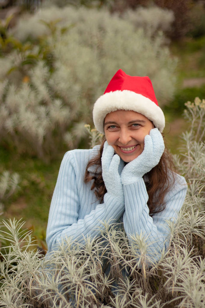 Young woman wearing red santa hat and blue gloves. Pretty Caucasian woman smiling and touching her face. Christmas and New Year holidays.  - Фото, зображення