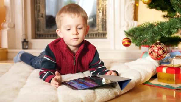 4k video of little boy lying on blanket under Christmas tree and playing video games on digital tablet computer. Child having fun on winter holidays and celebrations. - Materiał filmowy, wideo