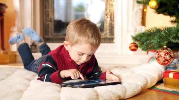 4k closeup video of little boy with digital tablet lying on floor at living room next to Christmas tree and fireplace. Child having fun on winter holidays and celebrations. - 映像、動画