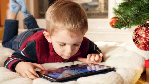 Nahaufnahme 4k Video eines lächelnden kleinen Jungen, der Spiele auf einem digitalen Tablet-Computer spielt, während er zu Hause unter dem Weihnachtsbaum liegt. Kinder haben Spaß beim Winterurlaub und Feiern. - Filmmaterial, Video