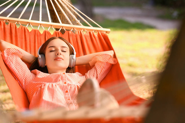 Όμορφη νεαρή γυναίκα με ακουστικά που ξεκουράζονται σε αιώρα σε εξωτερικούς χώρους - Φωτογραφία, εικόνα