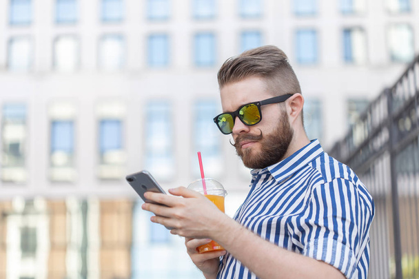 Πλευρική όψη ενός νεαρού κομψού χίπστερ με γυαλιά και ένα μουστάκι και χυμό στα χέρια του να συνομιλεί σε κοινωνικά δίκτυα χρησιμοποιώντας ένα smartphone και ασύρματο Internet σε μια ζεστή καλοκαιρινή μέρα. - Φωτογραφία, εικόνα