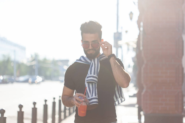 Portrait of a positive cheerful young arab man with a glass of juice with a straw while walking in the city on a warm sunny summer day. The concept of rest after study and work on weekends. - Foto, immagini