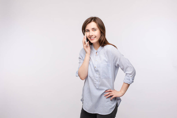 Smiling girl talking on the phone.Studio portrait of pretty young brunette woman in casual shirt and jeans talking on the mobile phone and smiling at camera. Isolate on white. - Foto, Imagen