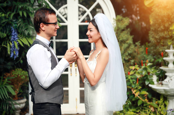 schöne kaukasische Bräutigam und asiatische Braut halten Hände und Augen Kontakt mit Lächeln und glückliches Gesicht während der Hochzeit oder vor der Hochzeit Foto-Pose im Garten  - Foto, Bild