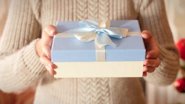 Closeup 4k footage of young woman holding beautiful box with gift tied with ribbon and bow. Family giving and receiving New Year presents in living room next to Christmas tree. - Footage, Video