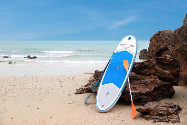 бело-голубая доска для серфинга на песчаном пляже с голубым морем и небом на фоне
 - Фото, изображение