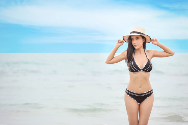 retrato de jovem asiático bonito e sexy mulher em preto biquíni e branco chapéu com beleza rosto de pé na praia arenosa com azul céu e esquerda cópia espaço - Foto, Imagem