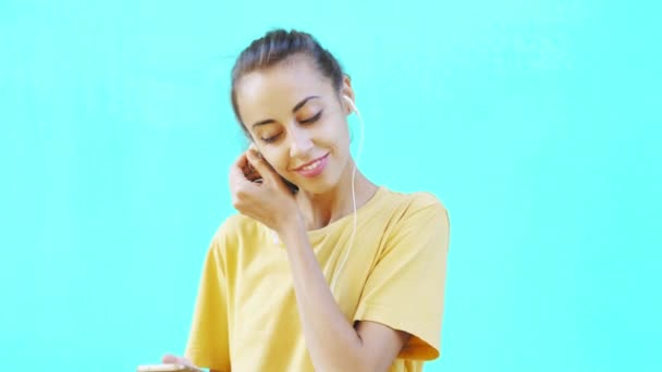 Pretty smiling young woman using smartphone and listening music with headphones, posing on a colorful bright cyan background. woman shows pleasure, enjoyment and positive emotions. - Filmati, video